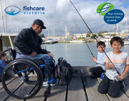 A man in a wheelchair and two young children beside him holding fishing rods on a jetty